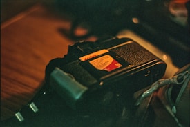 A vintage film camera lies on a wooden surface, partially in focus. The camera features a distinctive black textured body and a vibrant Kodak film label, suggesting analog photography. There are subtle highlights on metallic parts, indicating low ambient lighting.