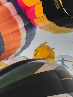 A vibrant gathering of hot air balloons fills the sky, featuring various colorful designs. In the foreground, a large balloon with a cartoonish yellow face adds a playful element. The different shapes and hues create a dynamic and festive atmosphere.