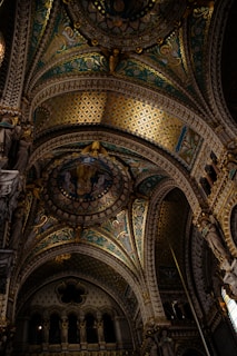 Intricate and ornate ceiling of a cathedral featuring detailed mosaics, arches, and architectural embellishments with rich colors and patterns. Statues adorn the sides, adding to the grandeur and solemnity.