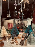 A window display features intricately designed paper ornaments, including stars and hanging decorations, alongside several paper-crafted Christmas trees in shades of green, teal, and beige. Below the trees, various boxes containing decorative items are arranged neatly on a draped white cloth. An angel figure made of paper stands to the right among the ornaments. Behind the decorations, signage with text is partially visible.