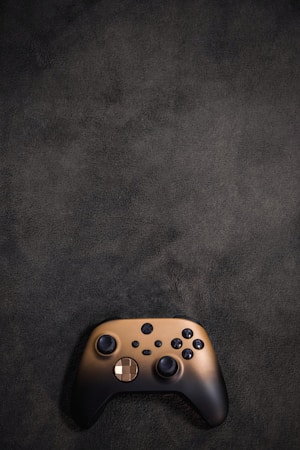 A black and gold video game controller placed on a dark textured surface. The controller has two joysticks, a D-pad, and several buttons, typical of modern gaming devices.