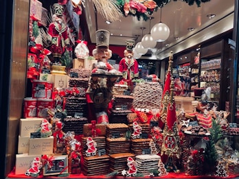 A festive bakery display with a variety of gingerbread and chocolate treats stacked in piles. Two large, decorative nutcracker figurines stand among the confections, adorned with red and gold regal costumes. The scene is accentuated with Christmas decorations, including small gift boxes, Santa figurines, and greenery.
