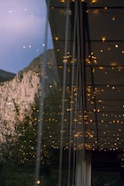 A view reflects a natural landscape with mountainous terrain and trees, seen through a glass panel. Attached to the panel are numerous small, warm-colored string lights, casting an inviting glow despite the cooler tones of the surroundings.