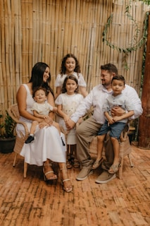 A family of six is gathered together, featuring three children and three adults. They are seated on wicker chairs set against a bamboo backdrop. The adults and children are dressed in coordinated light-colored clothing. The setting appears to be warm and inviting.