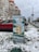 A utility box decorated with a mural stands in a snowy urban environment. The mural depicts a building and hot air balloons amidst greenery. In the background, there are several cars parked along a street and an apartment building with distinctive red-orange accents.