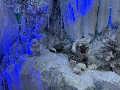 A winter-themed scene featuring a snow-covered environment. A lifelike snow leopard with a fluffy mane lies on icy terrain, surrounded by frost-covered trees and rocks. A white rabbit and a small bird are positioned nearby, adding to the naturalistic setting. The scene is illuminated with cool blue lighting, enhancing the icy atmosphere.