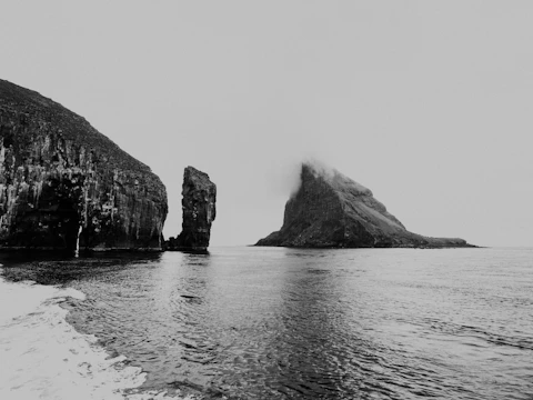 A black and white photograph of a coastal landscape featuring rugged cliffs and a prominent rock formation emerging from the water. The sky is overcast, adding a moody atmosphere to the scene. The water reflects the textures of the cliffs, creating a sense of tranquility.
