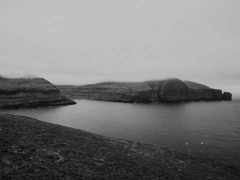 A black and white landscape depicting a large body of water surrounded by steep, rocky cliffs under a cloudy sky. The image has a calm and serene atmosphere with soft shadows and highlights.