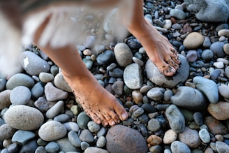 Bare feet are seen standing on a variety of smooth, rounded stones, with some sand adhering to the skin.