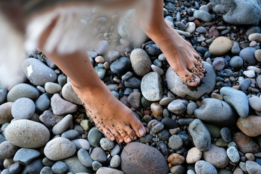Bare feet are seen standing on a variety of smooth, rounded stones, with some sand adhering to the skin.