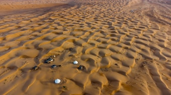 An expansive desert landscape filled with undulating sand dunes under the golden sunlight. Several small structures and tents are scattered across the area, indicating a temporary or makeshift settlement. The scene evokes a sense of isolation and vastness.