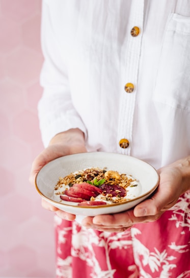 A person is holding a bowl of yogurt topped with granola, fresh fruit slices, and mint leaves. The individual is wearing a white shirt with brown buttons and a red patterned skirt. The background has a soft pink hexagonal pattern.