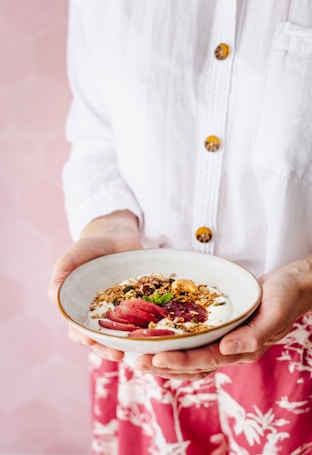A person is holding a bowl of yogurt topped with granola, fresh fruit slices, and mint leaves. The individual is wearing a white shirt with brown buttons and a red patterned skirt. The background has a soft pink hexagonal pattern.