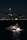 A yacht sails on a dark, calm body of water under a night sky. In the background, a distant city skyline is illuminated with lights, highlighting a tall tower with a bright red light at its peak.