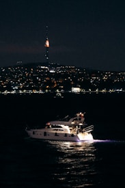 A yacht sails on a dark, calm body of water under a night sky. In the background, a distant city skyline is illuminated with lights, highlighting a tall tower with a bright red light at its peak.