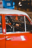 The interior of a vintage car with a red exterior and a steering wheel prominently visible. Hanging from the rearview mirror is an air freshener shaped like a tree with a crescent moon and star symbol. The dashboard features an old-fashioned radio. The background is blurred, showing some blue and warm lighting.
