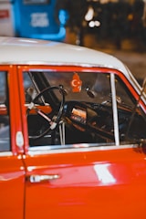 The interior of a vintage car with a red exterior and a steering wheel prominently visible. Hanging from the rearview mirror is an air freshener shaped like a tree with a crescent moon and star symbol. The dashboard features an old-fashioned radio. The background is blurred, showing some blue and warm lighting.