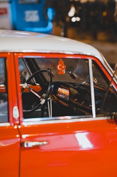 The interior of a vintage car with a red exterior and a steering wheel prominently visible. Hanging from the rearview mirror is an air freshener shaped like a tree with a crescent moon and star symbol. The dashboard features an old-fashioned radio. The background is blurred, showing some blue and warm lighting.