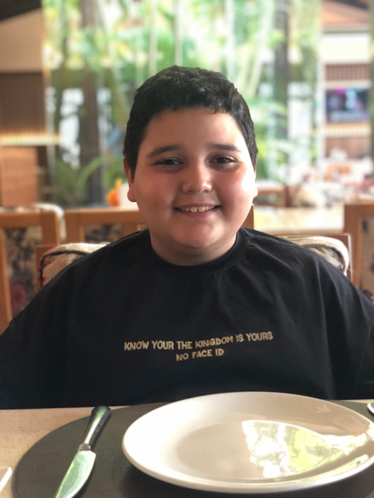 A smiling child is sitting at a dining table with an empty white plate and a knife placed on a black placemat. They are wearing a black T-shirt with a printed message that reads, 'KNOW YOUR THE KINGDOM IS YOURS NO FACE ID.' The background is blurred, showing greenery and a warm, indoor setting with wooden elements.