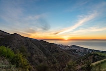 A picturesque landscape with the sun setting over the ocean, casting vibrant hues across the sky. Rolling hills are covered in greenery, and there is a coastal town nestled at the base of the hills near the water.