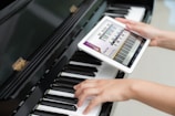 A person is holding a tablet displaying a musical scale app over a piano keyboard. One hand is placed on the piano keys, and the other hand is holding the tablet screen which shows a detailed musical notation.