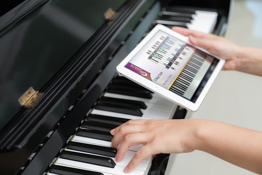 A person is holding a tablet displaying a musical scale app over a piano keyboard. One hand is placed on the piano keys, and the other hand is holding the tablet screen which shows a detailed musical notation.