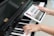 A person is holding a tablet displaying a musical scale app over a piano keyboard. One hand is placed on the piano keys, and the other hand is holding the tablet screen which shows a detailed musical notation.