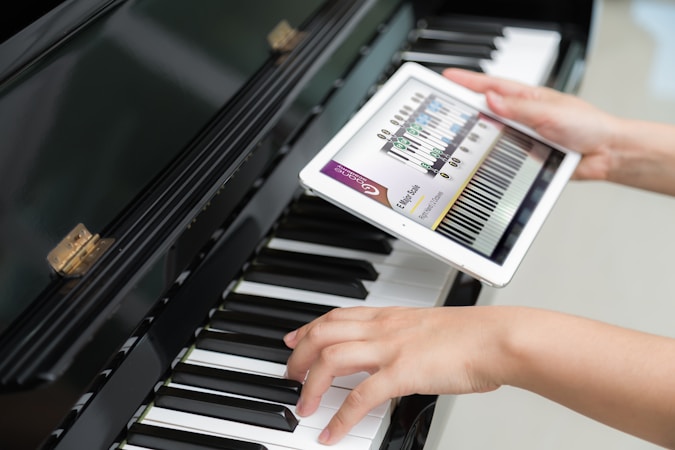 A person is holding a tablet displaying a musical scale app over a piano keyboard. One hand is placed on the piano keys, and the other hand is holding the tablet screen which shows a detailed musical notation.