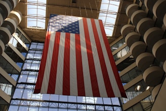 A large American flag hangs inside a modern building with a high ceiling and extensive glass windows. The flag's stripes are prominently displayed, and the building's architecture features symmetrical balconies and a skylight.