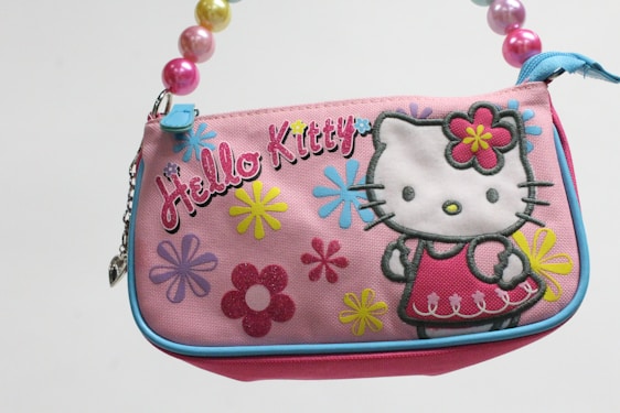 A colorful handbag featuring the character Hello Kitty, decorated with large floral patterns in various colors such as blue, yellow, and pink. The bag is primarily pink, with a blue trim around its edges. The handle is made up of large, shiny beads in different colors.
