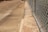 A concrete sidewalk runs parallel to a chain-link fence with metal posts. The ground appears weathered with some stains and patches. There are shadows and sunlight casting across the sidewalk and fence, suggesting it is an outdoor scene during the daytime.