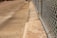 A concrete sidewalk runs parallel to a chain-link fence with metal posts. The ground appears weathered with some stains and patches. There are shadows and sunlight casting across the sidewalk and fence, suggesting it is an outdoor scene during the daytime.
