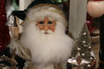 A close-up of a Santa Claus figure with a white beard and bushy eyebrows, wearing a green fur-lined hat and a coat. The figure holds a miniature Christmas tree decorated with silver ornaments. A small bell and a reindeer figure are also visible in the foreground.