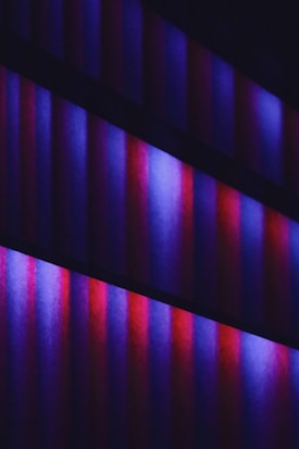 A series of vertical bars in gradient shades of purple, blue, and red arranged in parallel rows. The bars emanate a soft glowing effect against a dark background, creating a visually striking and modern aesthetic.