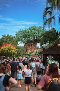 A large artificial tree with a thick trunk and lush green foliage stands prominently, surrounded by various other plants. A crowd of people is walking towards the tree, some wearing colorful clothing. The sky is bright blue with scattered clouds, and there are tropical elements such as palm leaves in the foreground.