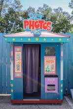 A colorful photo booth with a retro design. It features a bright blue exterior with red and yellow accents, covered in star motifs. There are two large posters on either side of the booth's entrance, advertising photo services. The entrance is partially covered by a purple curtain. Trees and a wooden fence are present in the background.