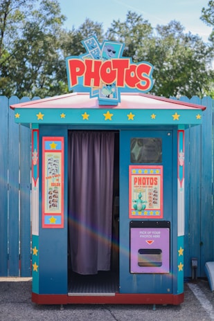 A colorful photo booth with a retro design. It features a bright blue exterior with red and yellow accents, covered in star motifs. There are two large posters on either side of the booth's entrance, advertising photo services. The entrance is partially covered by a purple curtain. Trees and a wooden fence are present in the background.