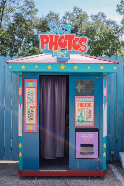 A colorful photo booth with a retro design. It features a bright blue exterior with red and yellow accents, covered in star motifs. There are two large posters on either side of the booth's entrance, advertising photo services. The entrance is partially covered by a purple curtain. Trees and a wooden fence are present in the background.