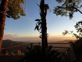 A scenic view of a wooden deck overlooking a mountainous landscape during sunset. The sky is filled with warm hues as the sun sets, and lush trees frame the deck. The horizon mixes mountains and clouds, creating a tranquil and picturesque atmosphere.