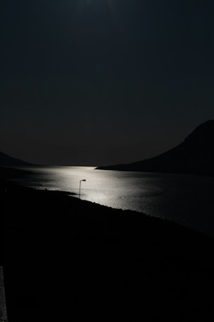 A serene night scene featuring a body of water reflecting the moonlight, flanked by dark silhouettes of mountains. A solitary lamp post stands on the edge of the shoreline.