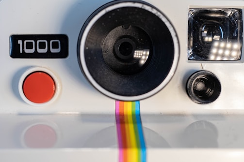 A vintage instant camera with a prominent black lens, a red shutter button, and a rainbow stripe running down the front. The camera displays the number '1000' and has a classic design with a simple, retro aesthetic.