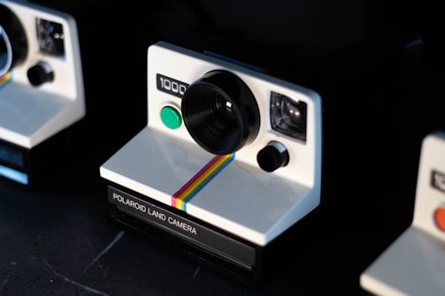 A close-up view of vintage Polaroid instant cameras displaying their retro design with a white body, black lens, and a colorful rainbow stripe accent. The cameras are positioned on a dark surface, suggesting a classic and nostalgic theme.