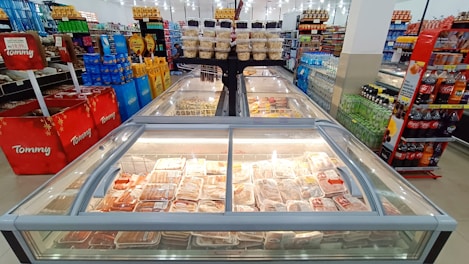 A supermarket aisle features rows of frozen food and refrigerated items in large glass cases. Shelves are stocked with a variety of beverages, snacks, and packaged goods. Bright lighting overhead enhances the visibility of products. The setting is orderly and well-organized, showcasing a typical grocery shopping environment.