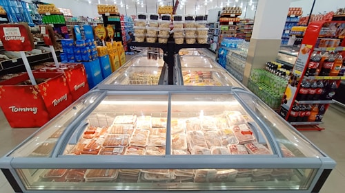 A supermarket aisle features rows of frozen food and refrigerated items in large glass cases. Shelves are stocked with a variety of beverages, snacks, and packaged goods. Bright lighting overhead enhances the visibility of products. The setting is orderly and well-organized, showcasing a typical grocery shopping environment.