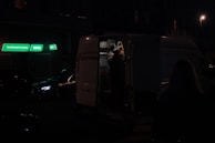 A dimly lit street scene featuring a parked van with its back doors open. The ambient lighting is low, with a few neon signs in green providing some illumination. A person is visible inside the van, reaching or organizing something. Nearby, another car is parked on the street.