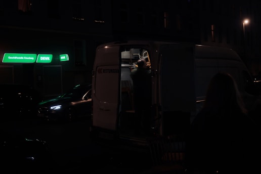 A dimly lit street scene featuring a parked van with its back doors open. The ambient lighting is low, with a few neon signs in green providing some illumination. A person is visible inside the van, reaching or organizing something. Nearby, another car is parked on the street.