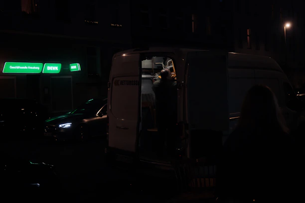 A nighttime roadside scene with a mobile mechanic's van parked next to a customer's vehicle under soft street lighting.