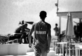 A lifeguard stands with the word 'SALVAMENTO' on the back of their clothing. Other people are visible, including someone lounging and a person standing near a structure. The setting appears to be an outdoor area, possibly a beach or poolside, with umbrellas and lounge chairs.