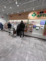 Several people are standing at a pharmacy counter with a selection of medications displayed behind a clear glass partition. The store has a modern design with bright lighting and digital displays showing numbers above each counter.