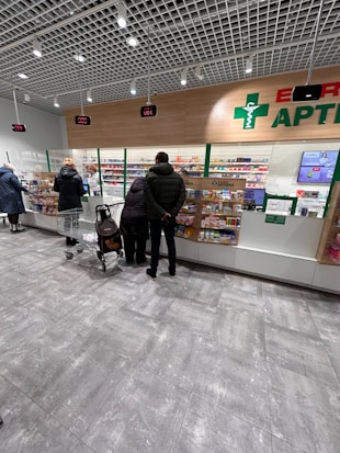 Several people are standing at a pharmacy counter with a selection of medications displayed behind a clear glass partition. The store has a modern design with bright lighting and digital displays showing numbers above each counter.